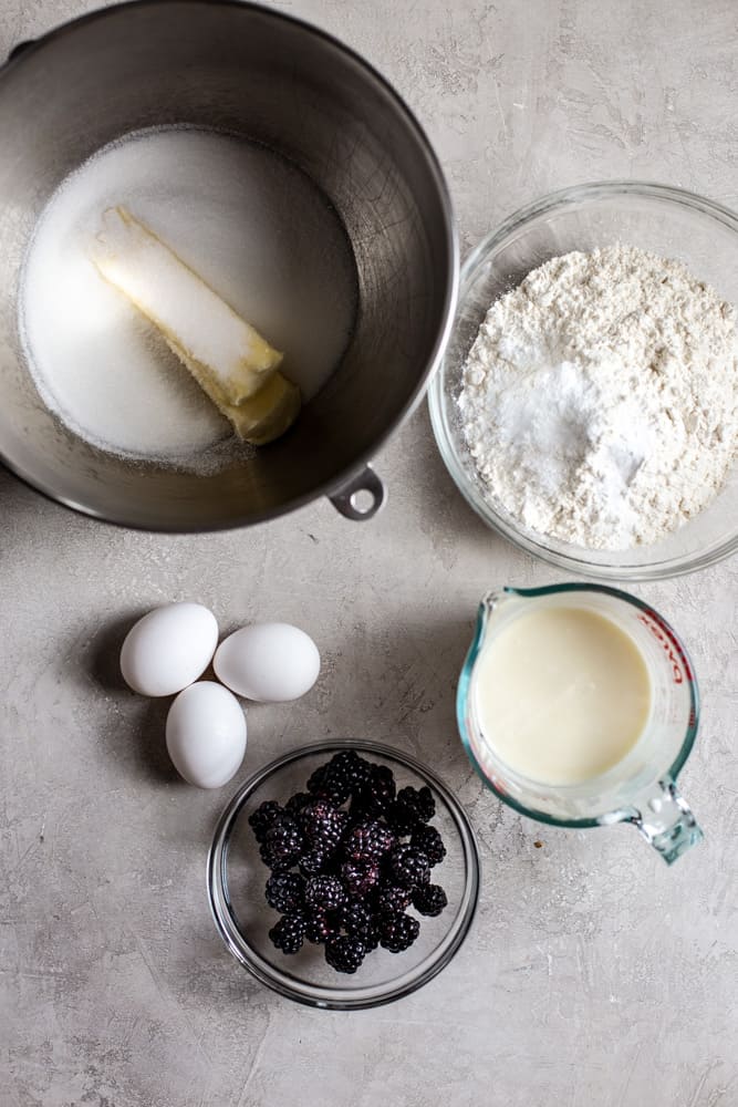 A mixing bowl with butter and sugar on a gray background next to a bowl of flour, 3 eggs, a measuring cup with milk and a bowl of blackberries