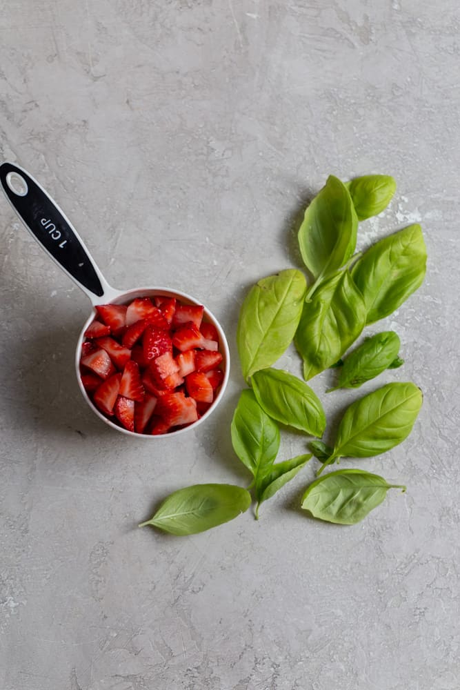 Chopped strawberries in a measuring cup next to loose basil on a gray background.