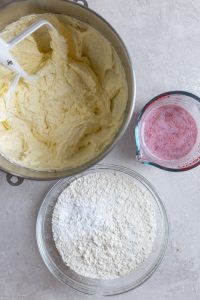 Cake batter in a mixing bowl next to bowl of flour and a mixture of milk and strawberries on a gray background