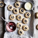 Glazed Raspberry thumbprint cookies on a parchment lined baking tray next to a bowl of jam