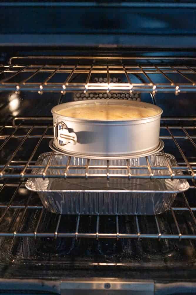 A cheesecake in the oven with a tray of water underneath it on the bottom rack.