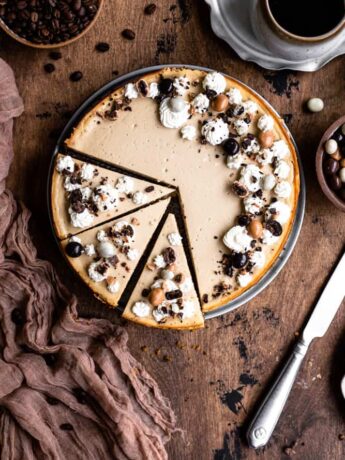 A cheesecake decorated with whipped cream and chocolate covered espresso beans with 3 slices cut on a wooden surface