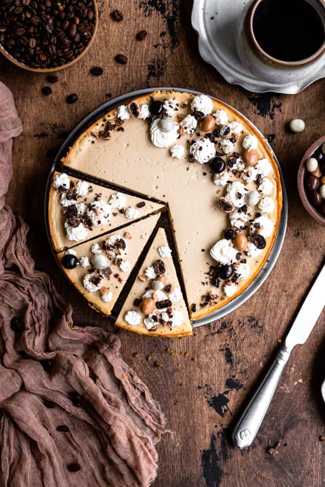 A cheesecake decorated with whipped cream and chocolate covered espresso beans with 3 slices cut on a wooden surface