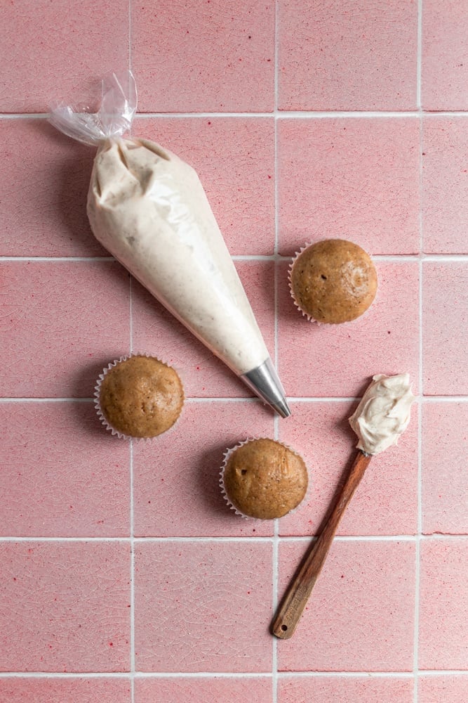 A piping bag filled with espresso buttercream next to 3 cupcakes on a pink tiled surface.