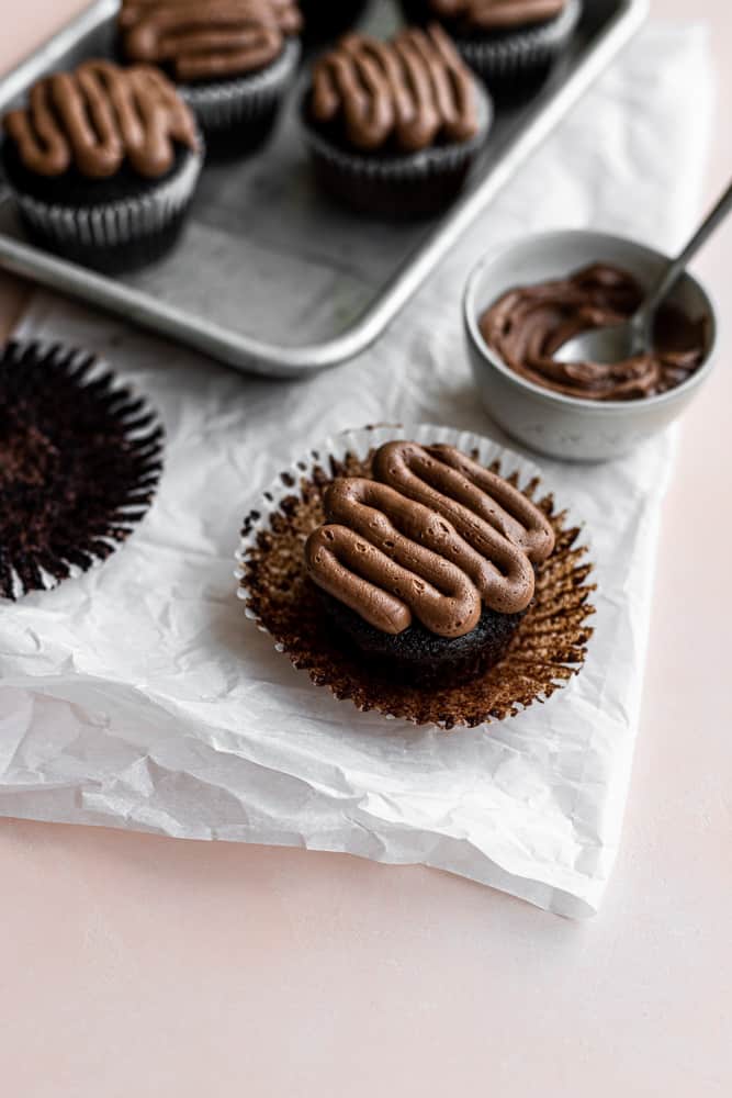 A chocolate cupcake with its liner pulled out on a piece of parchment paper.