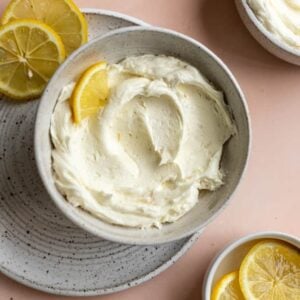 Lemon buttercream in a white bowl on a pink surface with a lemon slice in it.