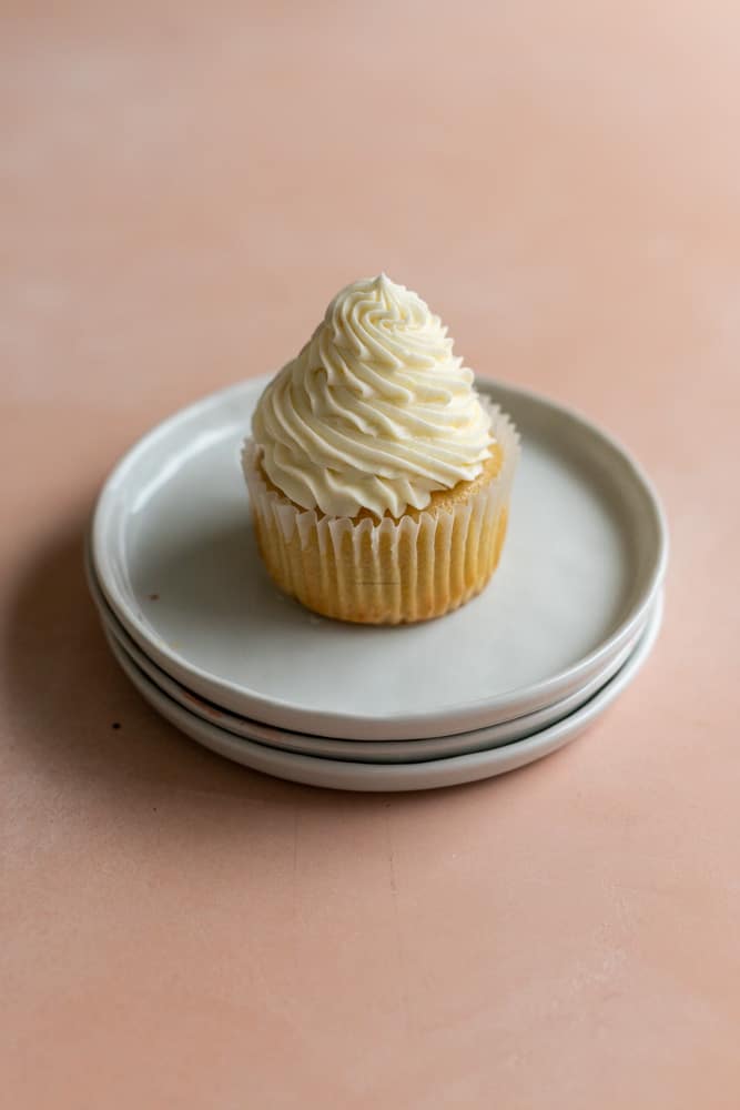 A vanilla cupcake with white icing on a stack of white plates on a pink background