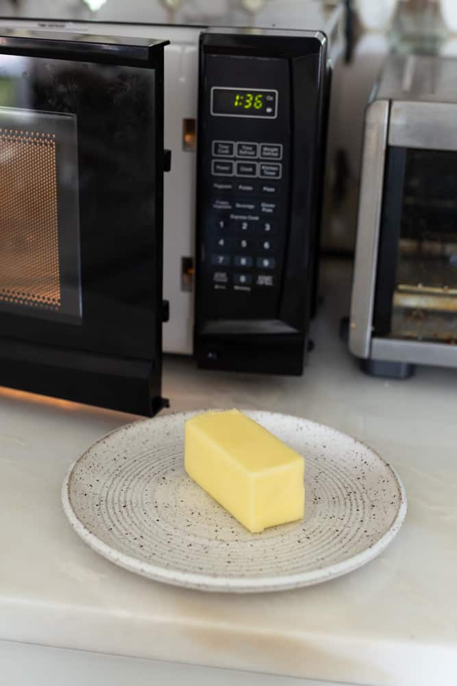 A stick of butter on a white plate next to a microwave with the door open