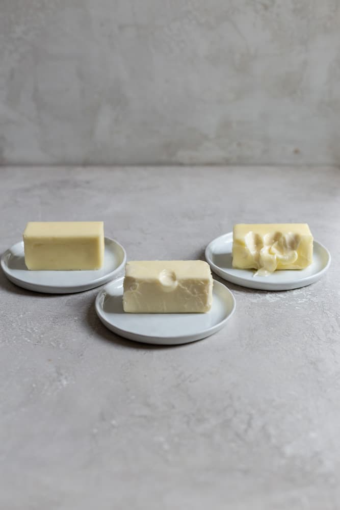 3 sticks of butter sitting on 3 plates on a gray surface