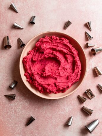 Bright pink raspberry frosting in a bowl on a pink surface surrounded by piping tips