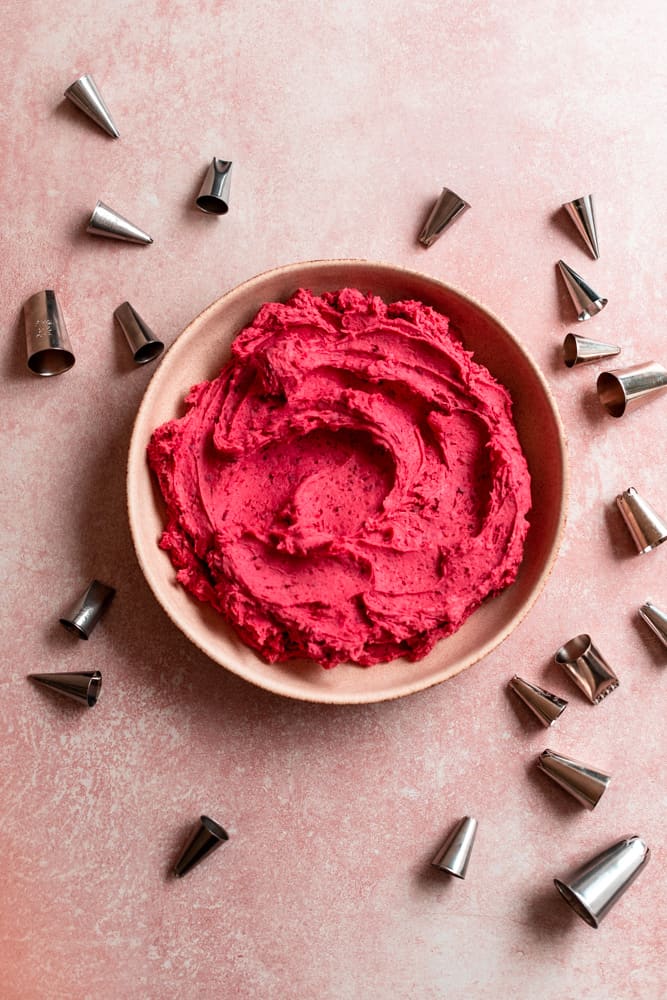 Bright pink raspberry frosting in a bowl on a pink surface surrounded by piping tips