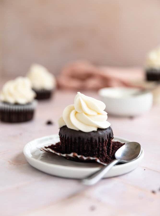 A chocolate cupcake with vanilla frosting on a white plate with a spoon next to it.