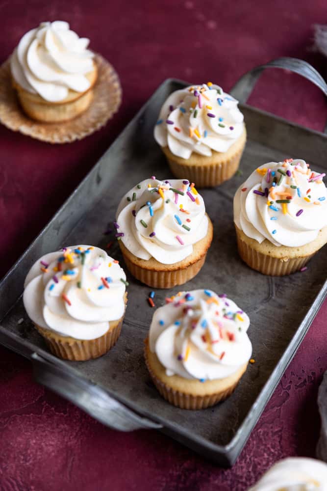 5 vanilla cupcakes with vanilla frosting and sprinkles in a tin on a deep red surface.