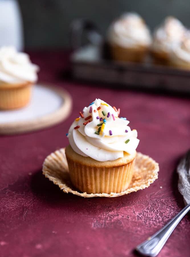 A vanilla cupcake topped with vanilla buttercream and sprinkles on a deep red surface.