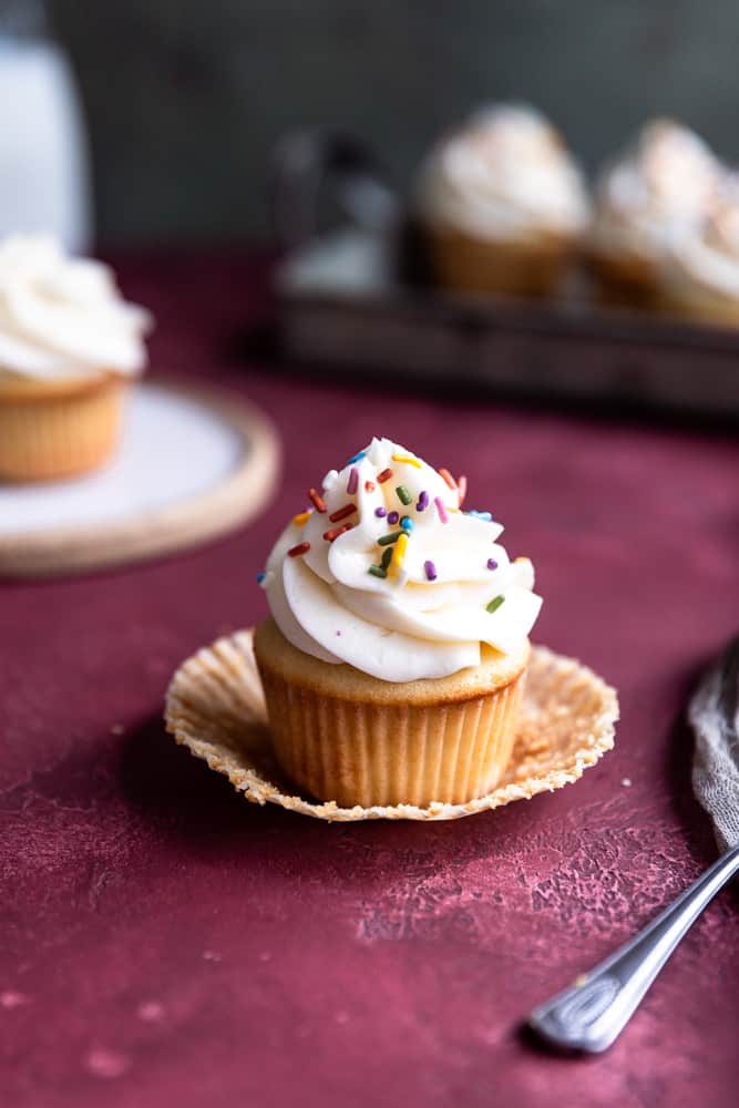 A vanilla cupcake topped with vanilla buttercream and sprinkles on a deep red surface.