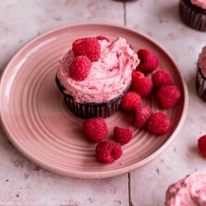 A chocolate raspberry cupcake on a pink plate with raspberries next to it.