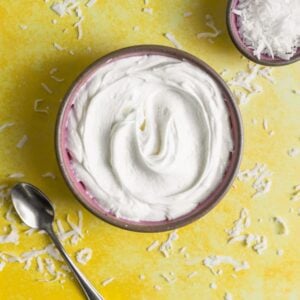 An overhead view of white frosting in a pink bowl on a yellow surface surrounded by shredded coconut.