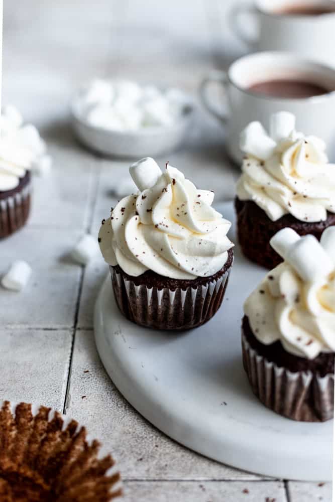 Hot chocolate cupcakes on a white plate with hot cocoa in the background.