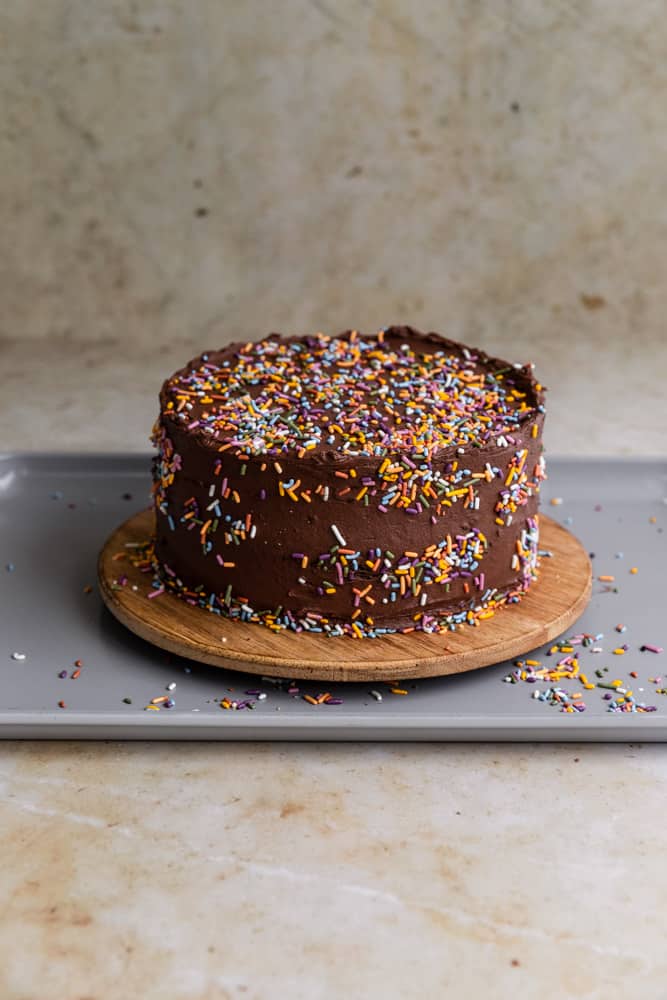 A chocolate cake covered in sprinkles sitting on a wooden serving board and sheet tray to catch loose sprinkles.