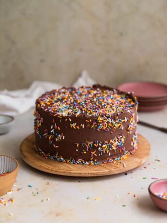 A chocolate cake covered in rainbow sprinkles on a wooden round serving tray.