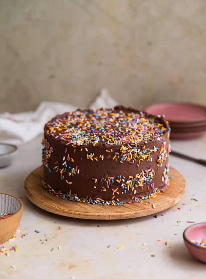 A chocolate cake covered in rainbow sprinkles on a wooden round serving tray.