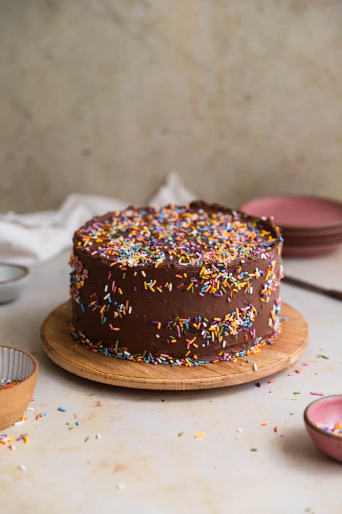A chocolate cake covered in rainbow sprinkles on a wooden round serving tray. 