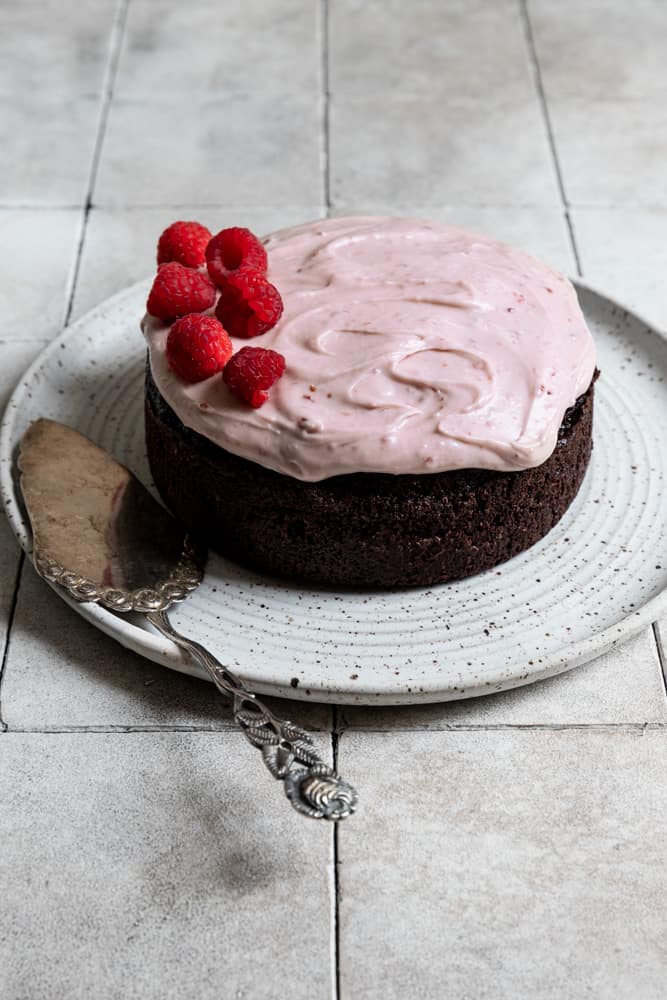 A chocolate cake topped with raspberry cream cheese frosting and fresh raspberries on a white plate.