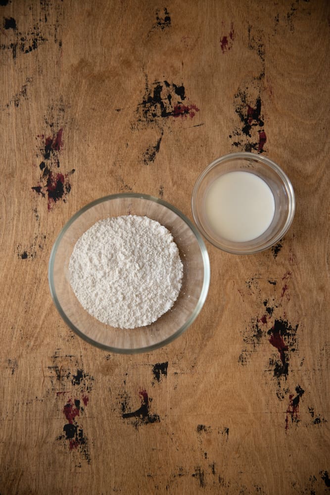 Confectioners sugar and milk in bowls on a wooden surface.
