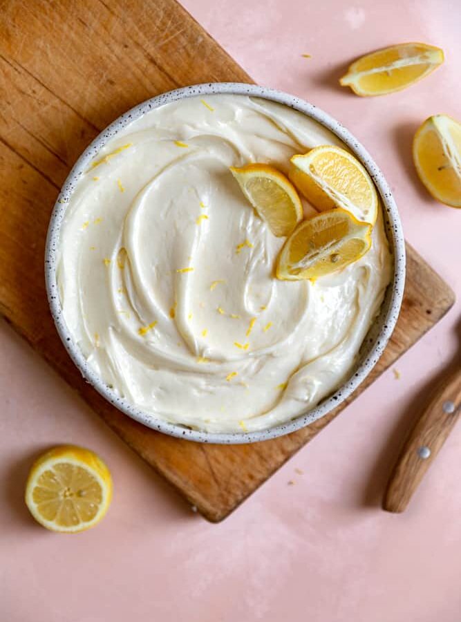 Lemon cream cheese frosting in a bowl with lemon slices on top sitting on a wooden cutting board.