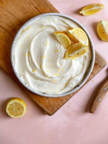 Lemon cream cheese frosting in a bowl with lemon slices on top sitting on a wooden cutting board.