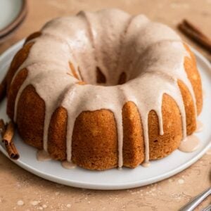 A cinnamon bundt cake topped with glaze on a white plate on a beige surface.