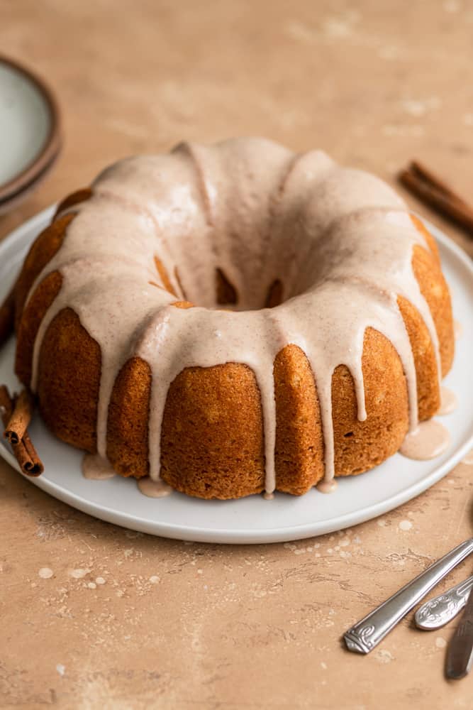 A cinnamon bundt cake topped with glaze on a white plate on a beige surface.