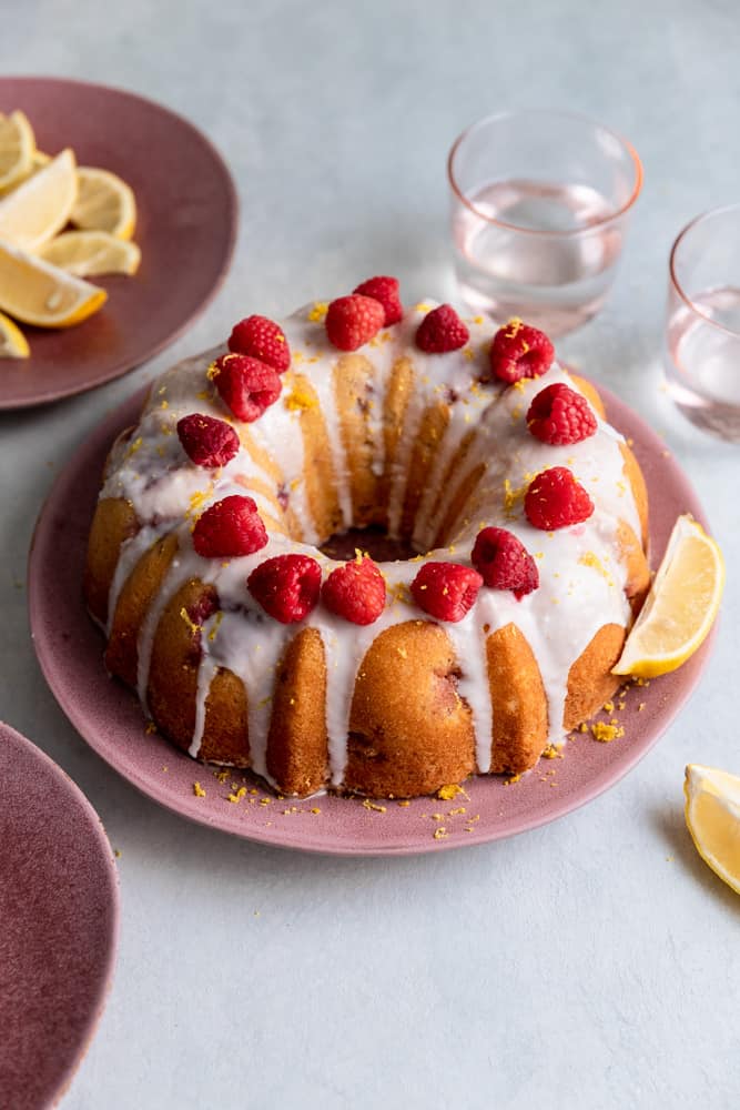 A bundt cake topped with white glaze, raspberries and lemon on a pink plate.