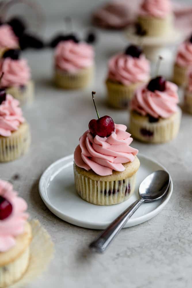 A cherry cupcake with pink frosting on a white small plate.