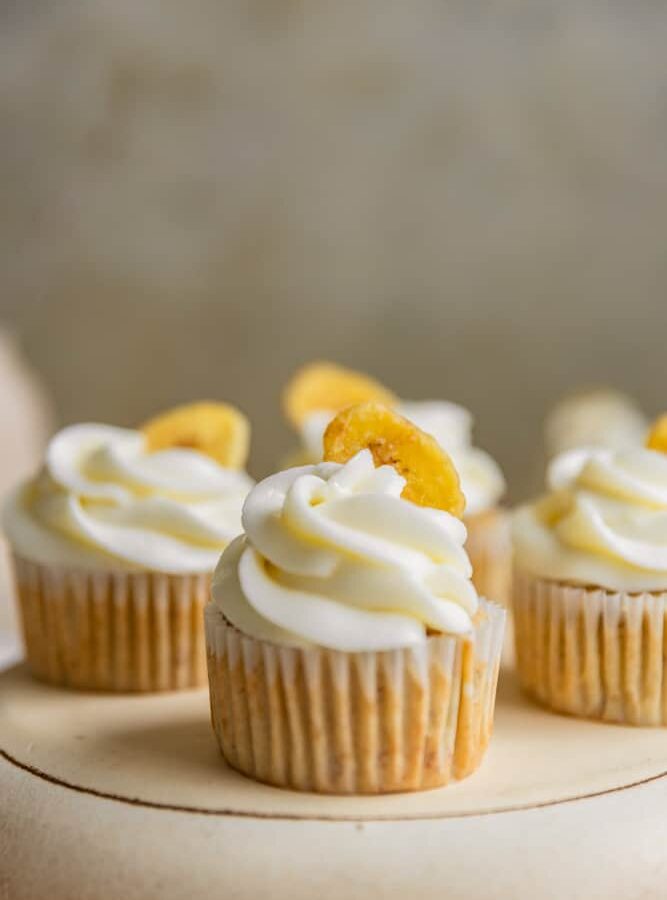 A closeup of a banana cupcake frosted with cream cheese frosting and garnished with a banana chip.