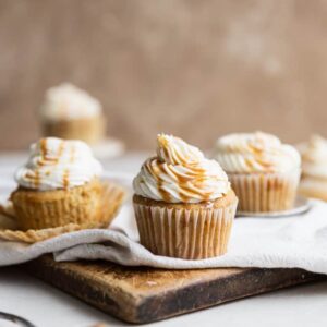 Caramel cupcakes with caramel drizzle on a linen and wooden cutting board.