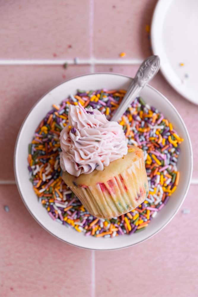 One confetti cupcakes on its side in a bowl of sprinkles.