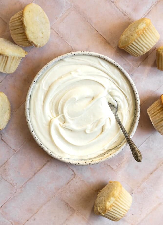Cream cheese frosting in a bowl next to unfrosted cupcakes on a pink tiled surface.