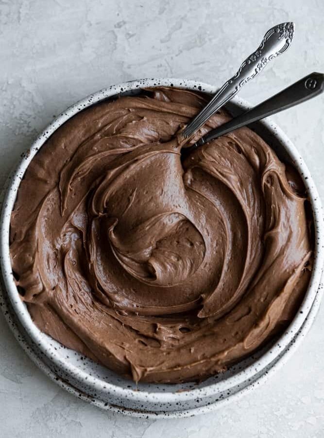 Chocolate cream cheese frosting swirled in a white bowl with 2 spoons.