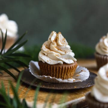 A gingerbread cupcake with its wrapper unwrapped on a wooden cutting board surrounded by greenery.