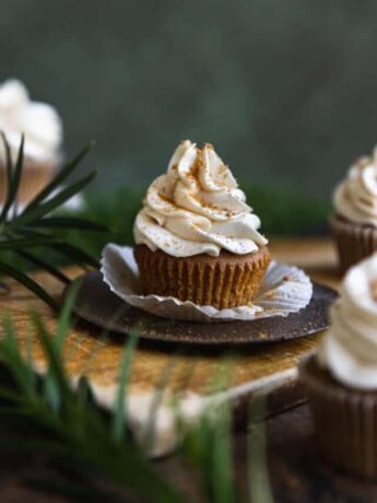 A gingerbread cupcake with its wrapper unwrapped on a wooden cutting board surrounded by greenery.