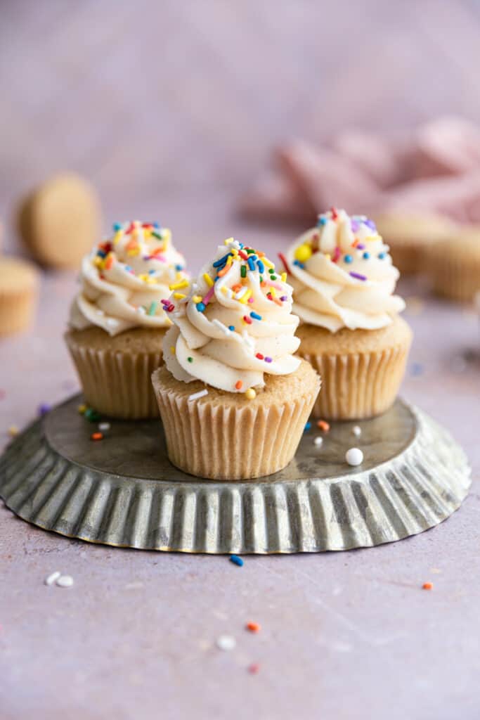 3 Vanilla cupcakes on an upside down tin frosted with buttercream and sprinkles.