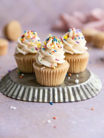 3 Vanilla cupcakes on an upside down tin frosted with buttercream and sprinkles.