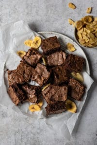 Banana brownie squares arranged on a plate next to banana chips.