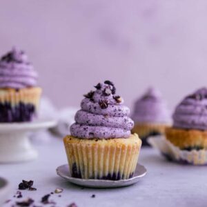 A blueberry cupcake with purple frosting on a tiny plate surrounded by other cupcakes.