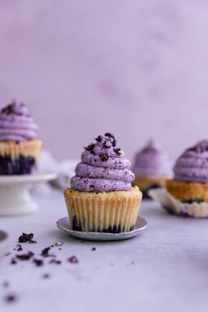 A blueberry cupcake with purple frosting on a tiny plate surrounded by other cupcakes.