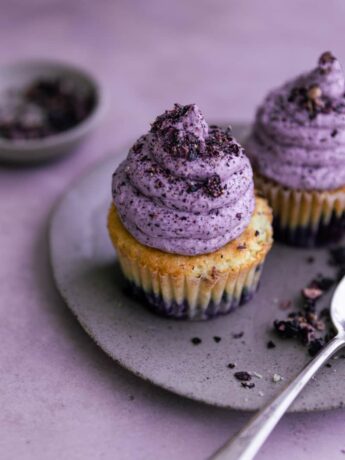 Two blueberry cupcakes on a plate on a purple surface.