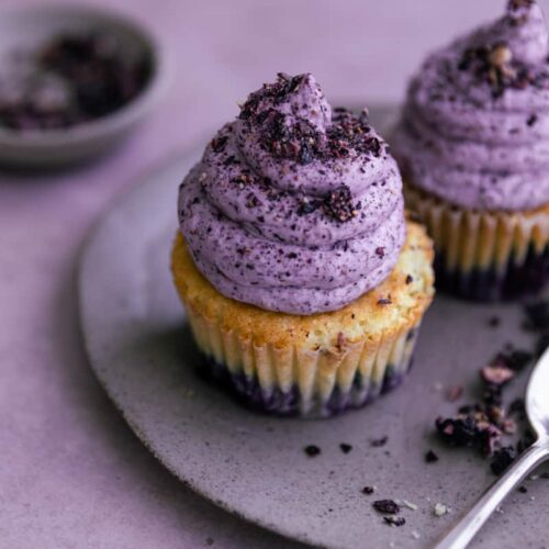 Two blueberry cupcakes on a plate on a purple surface.