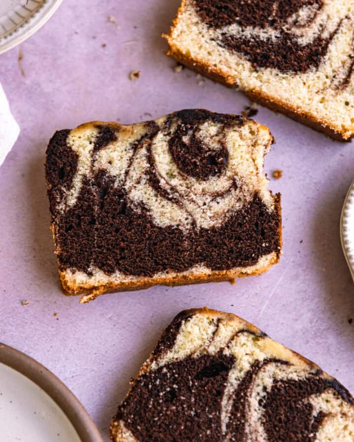 A slice of marble loaf cake next to a plate on a purple background.