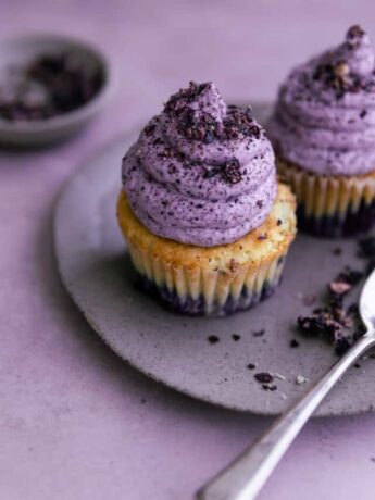 Two blueberry cupcakes on a plate on a purple surface.